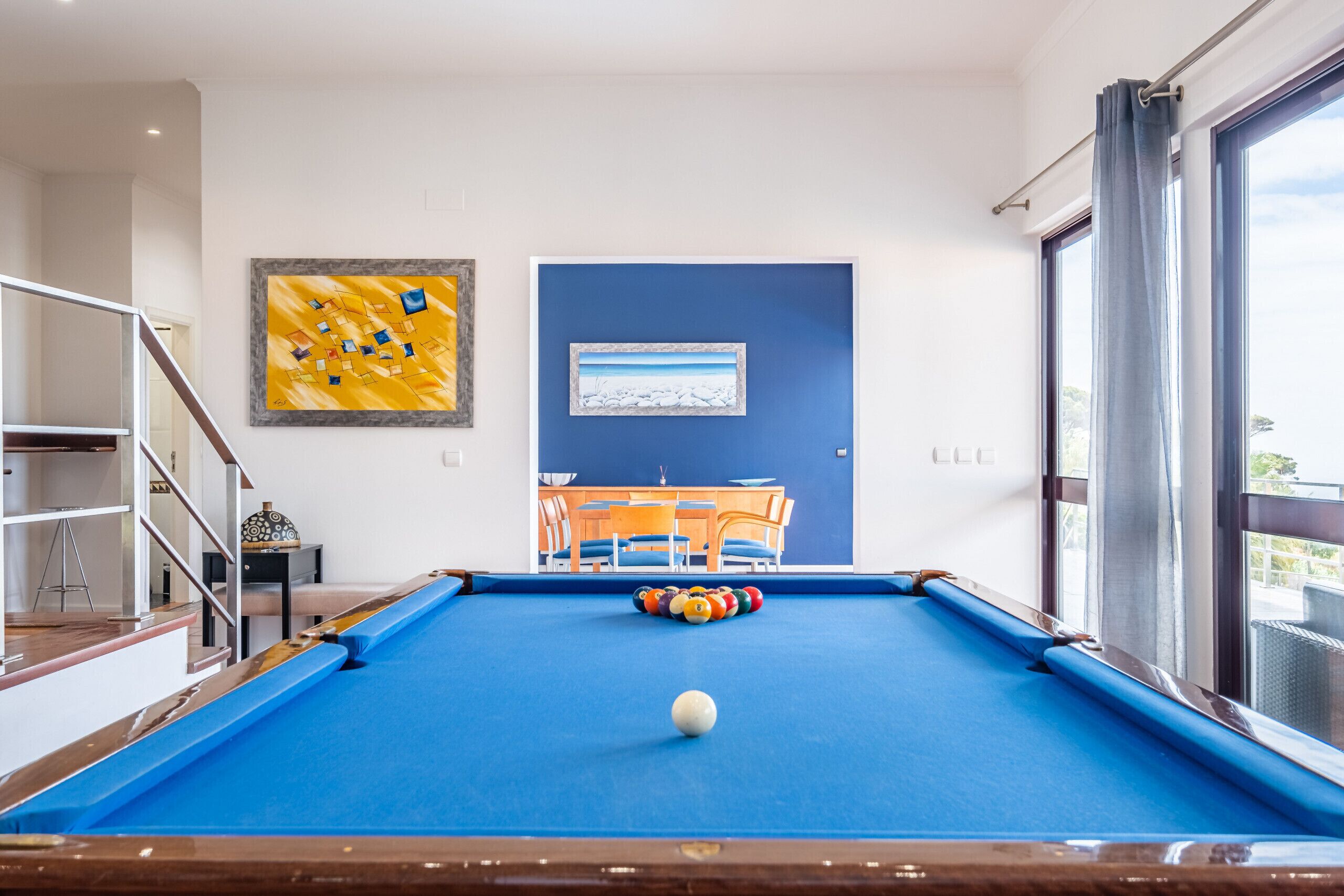 A pool table with a white cue ball, a dining area with a blue table and chairs, and a painting on the wall.