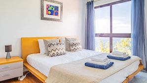A modern bedroom with a wooden headboard, a large window with curtains, a bedside table with a lamp, and a framed picture on the wall. - Vila Crerital - By Wehost (Funchal)
