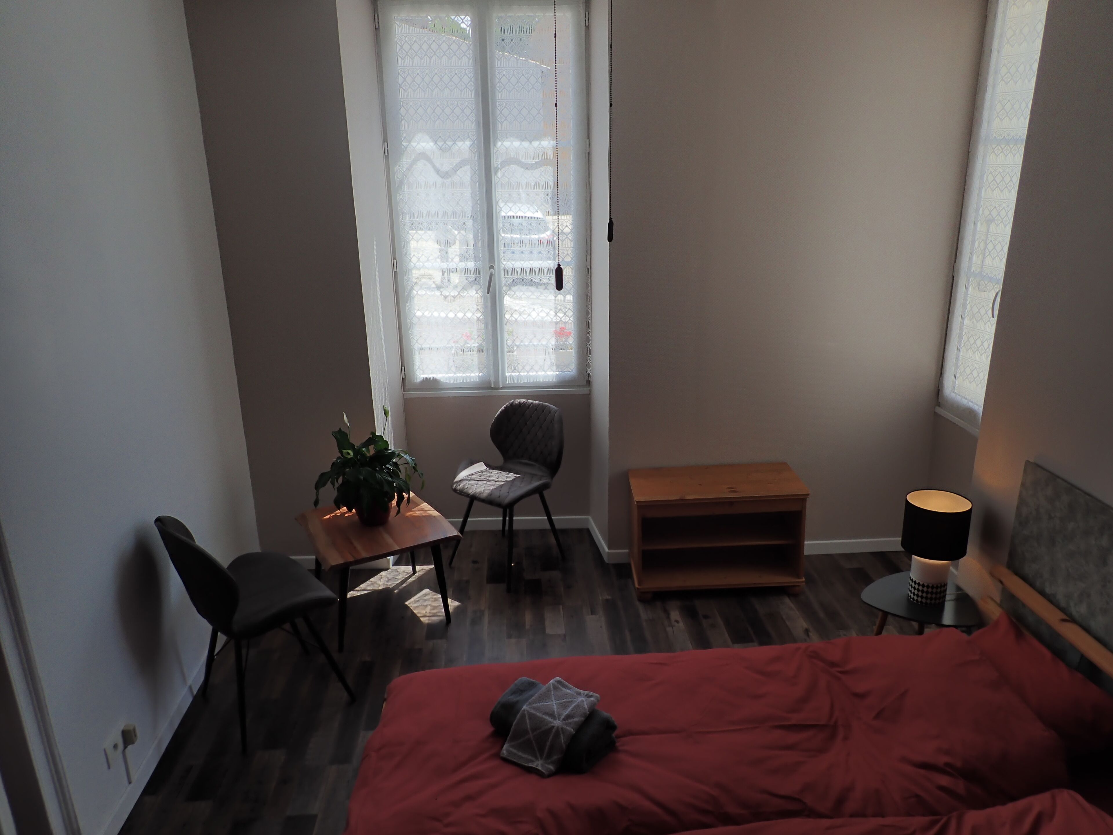 Un dormitorio pequeño y moderno con colcha roja, mesita de noche de madera, una silla, una mesita con una planta y una ventana con cortinas estampadas.