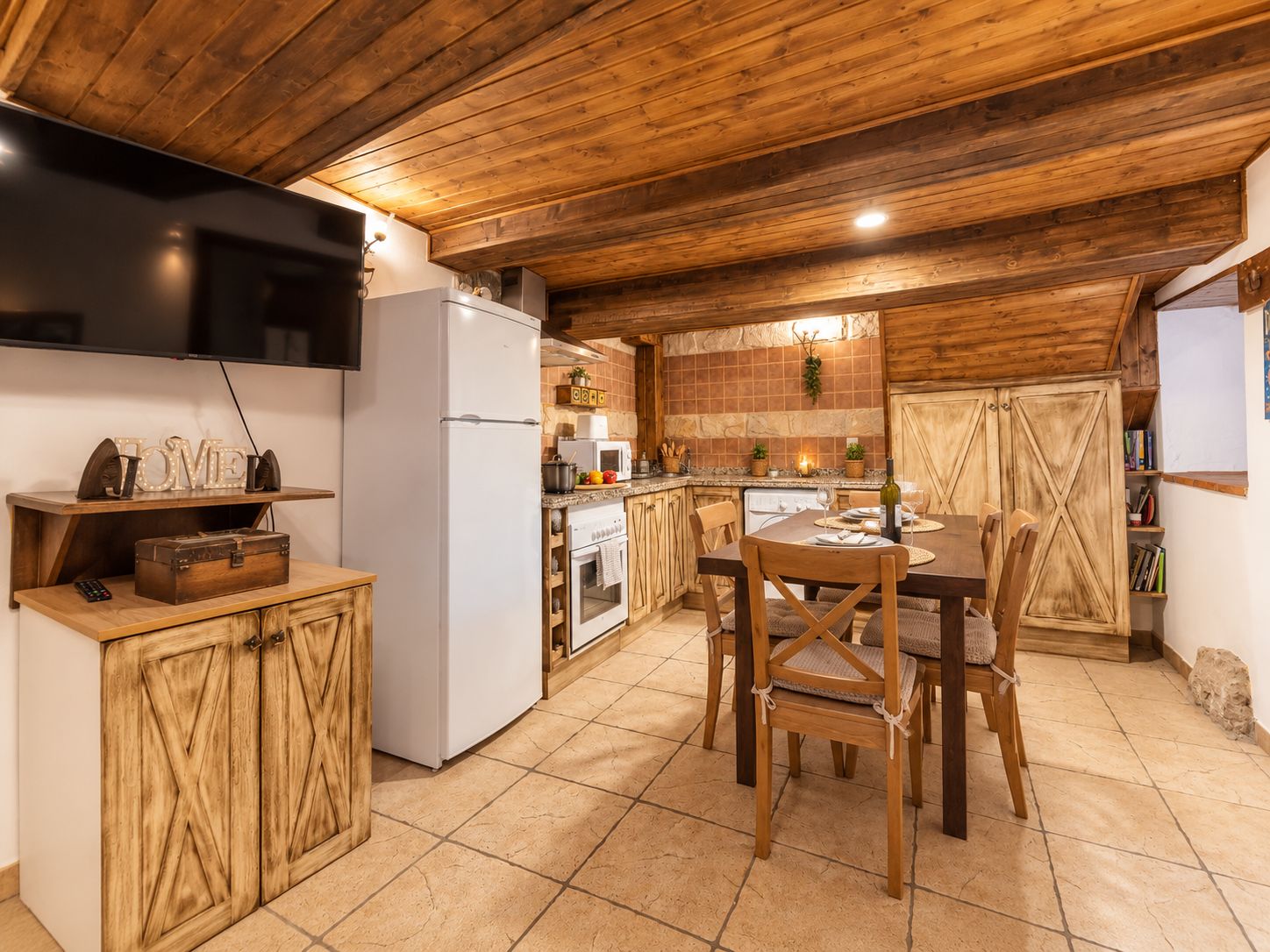 A rustic kitchen with wooden cabinets, a dining table with chairs, and a television on the wall.