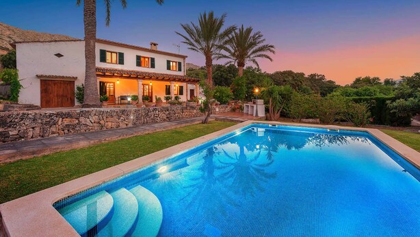 Una casa de dos pisos con piscina, muro de piedra y palmeras.