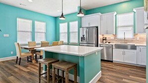 Una cocina con paredes azul verdoso, armarios blancos, una isla central con taburetes y un comedor con sillas estampadas.
