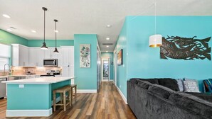 Una cocina moderna con armarios blancos, una isla central e iluminación colgante. Un pasillo conduce a una sala de estar con un sofá oscuro y paredes azul verdosas.