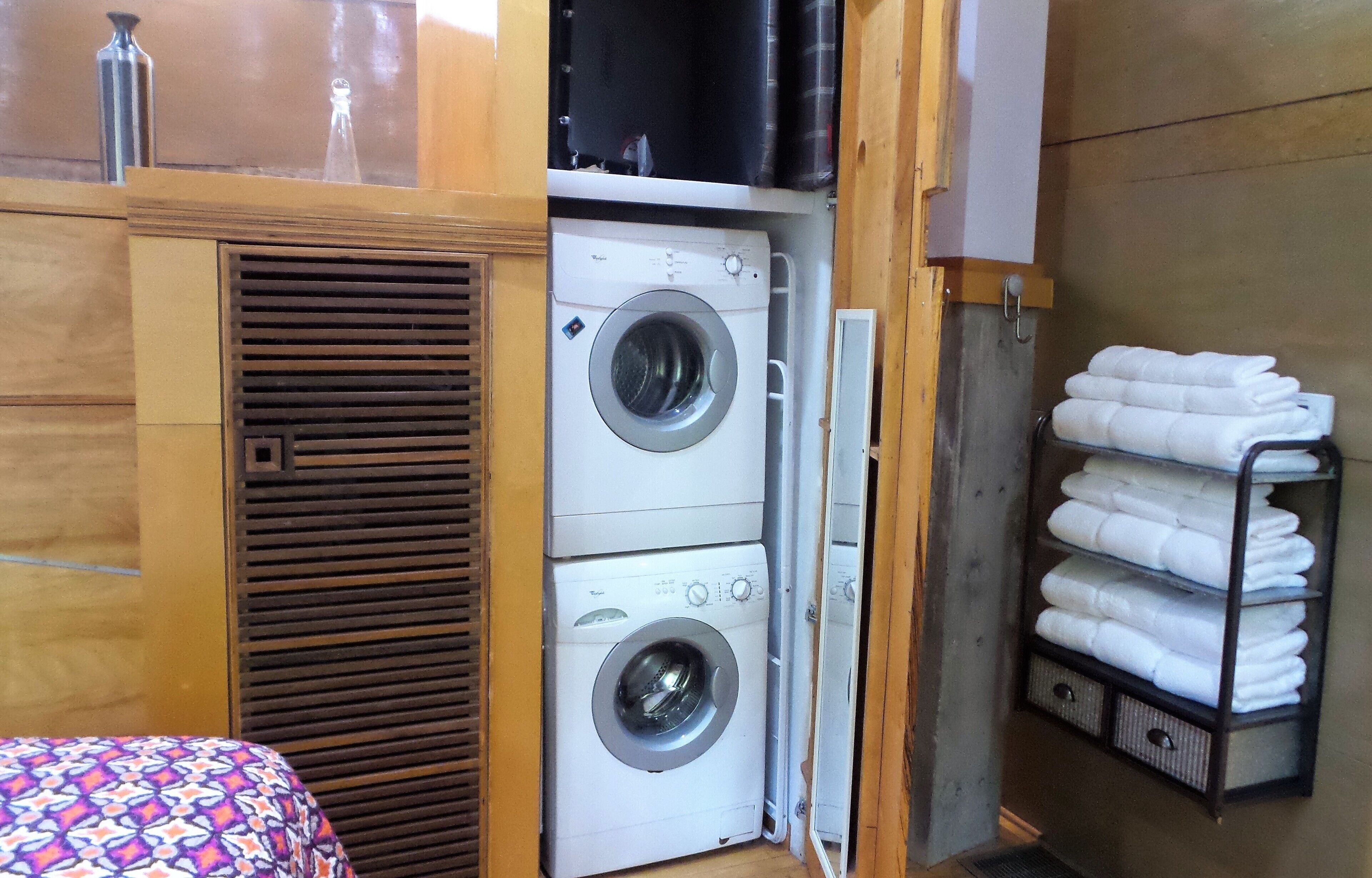 A compact room with a washing machine, a bed with a patterned bedspread, and a shelf with folded towels.