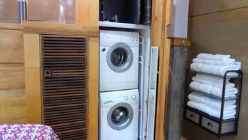 A compact room with a washing machine, a bed with a patterned bedspread, and a shelf with folded towels.