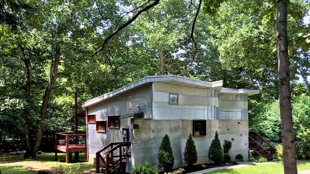 A small, single-story cabin with a metal exterior, surrounded by trees and a wooden deck. - One of a kind cabin in the woods.  Quiet and private but close to everything.  (Coxs Creek)