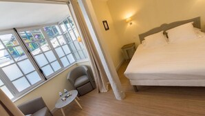 Habitación de hotel con una cama grande, una mesita pequeña con dos sillas, una ventana con vistas a edificios y una mesita de noche con una lámpara.
