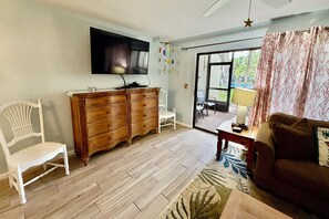 Una sala de estar con piso de madera, una silla blanca, un mueble de madera, un televisor y vistas al exterior a través de un ventanal.