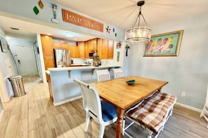 A kitchen with wooden cabinets, a stainless steel refrigerator, and a dining area with a wooden table and white chairs. - Updated 2 bedroom/2.5 bath townhouse, perfect for a large family beach getaway!! (Saint Augustine)