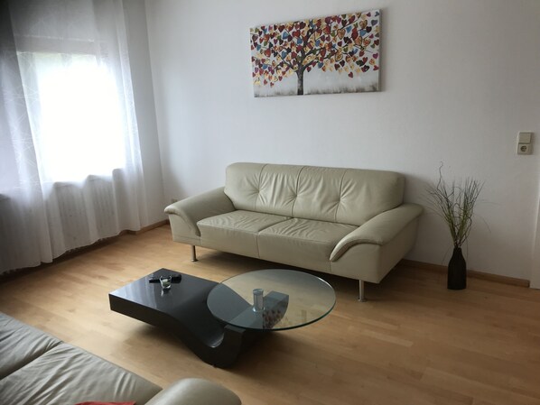 Een woonkamer met een beige bank, een glazen salontafel, een schilderij aan de muur en een vaas met planten.
