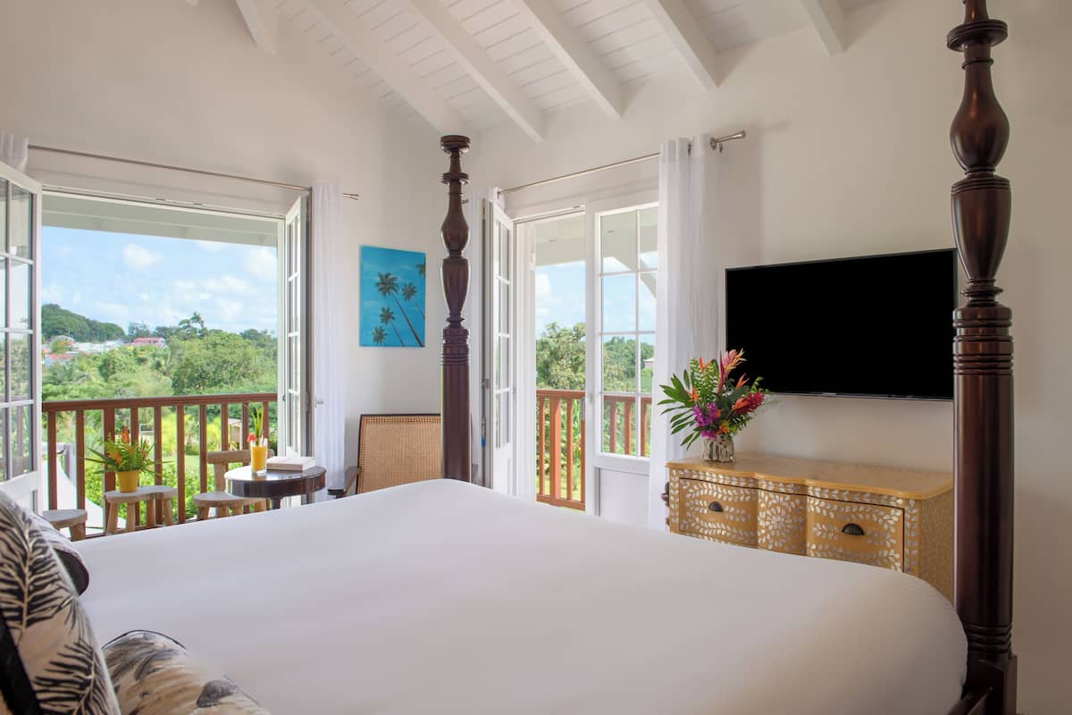 A bedroom with a large bed, a TV mounted on the wall, and a balcony with a view of the greenery.