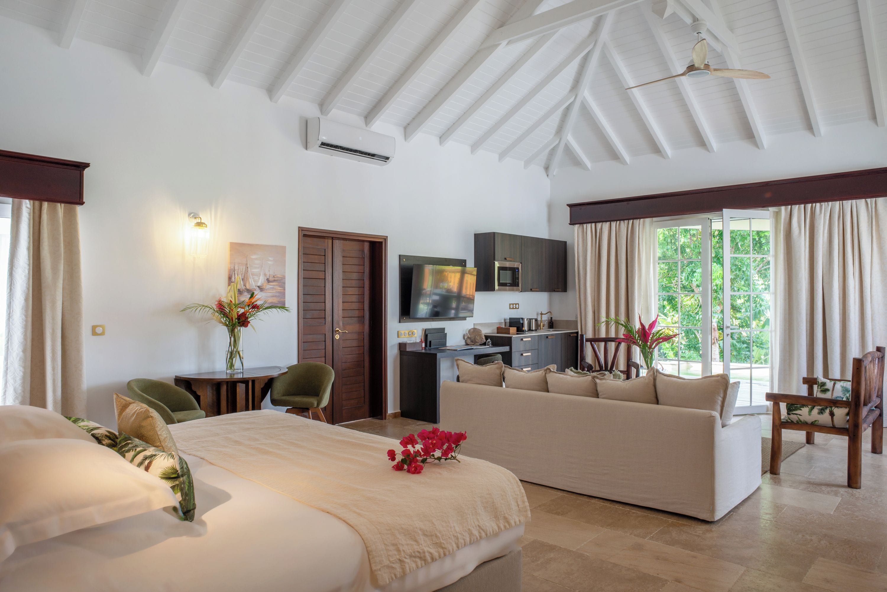 A hotel room with a bed, a sofa, a desk with a TV, a kitchenette, and a large window with curtains.