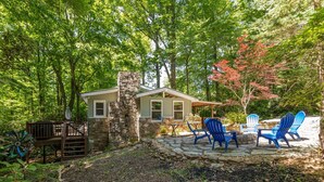 A rustic cabin with a stone chimney, surrounded by a forest. There are two Adirondack chairs and a wooden deck. - Cozy Cottage - Located on App Ski Mtn Lower level game room, Firepit (Blowing Rock)