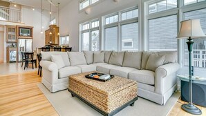 Een ruime woonkamer met een grote zithoekbank, een geweven salontafel, en een eethoek met stoelen, met een keuken op de achtergrond.