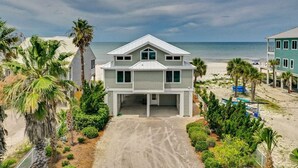 Een twee verdiepingen tellend strandhuis met een overdekte garage, omgeven door palmbomen en een zandpad.
