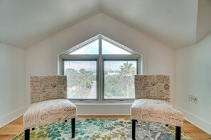 A room with a sloped ceiling, two chairs with vintage-style upholstery, a large window with a view of palm trees, and a patterned rug on the floor.