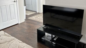 Una sala de estar con un televisor de pantalla plana sobre un soporte negro, una puerta blanca y un área de lavandería al fondo.