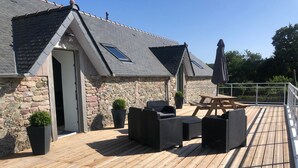 A wooden deck with outdoor furniture, a stone wall, and a building with a sloped roof. - Le domaine de frehel (Frehel)