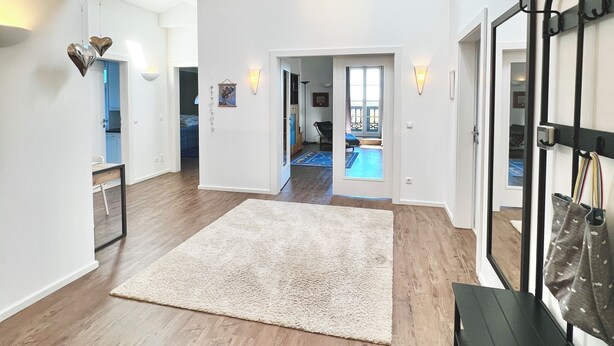 Ett rymligt rum med ett stort trägolv, en vit matta och en bänk. Det finns ett takfönster ovanför och genom en öppen dörr kan man se en pool.