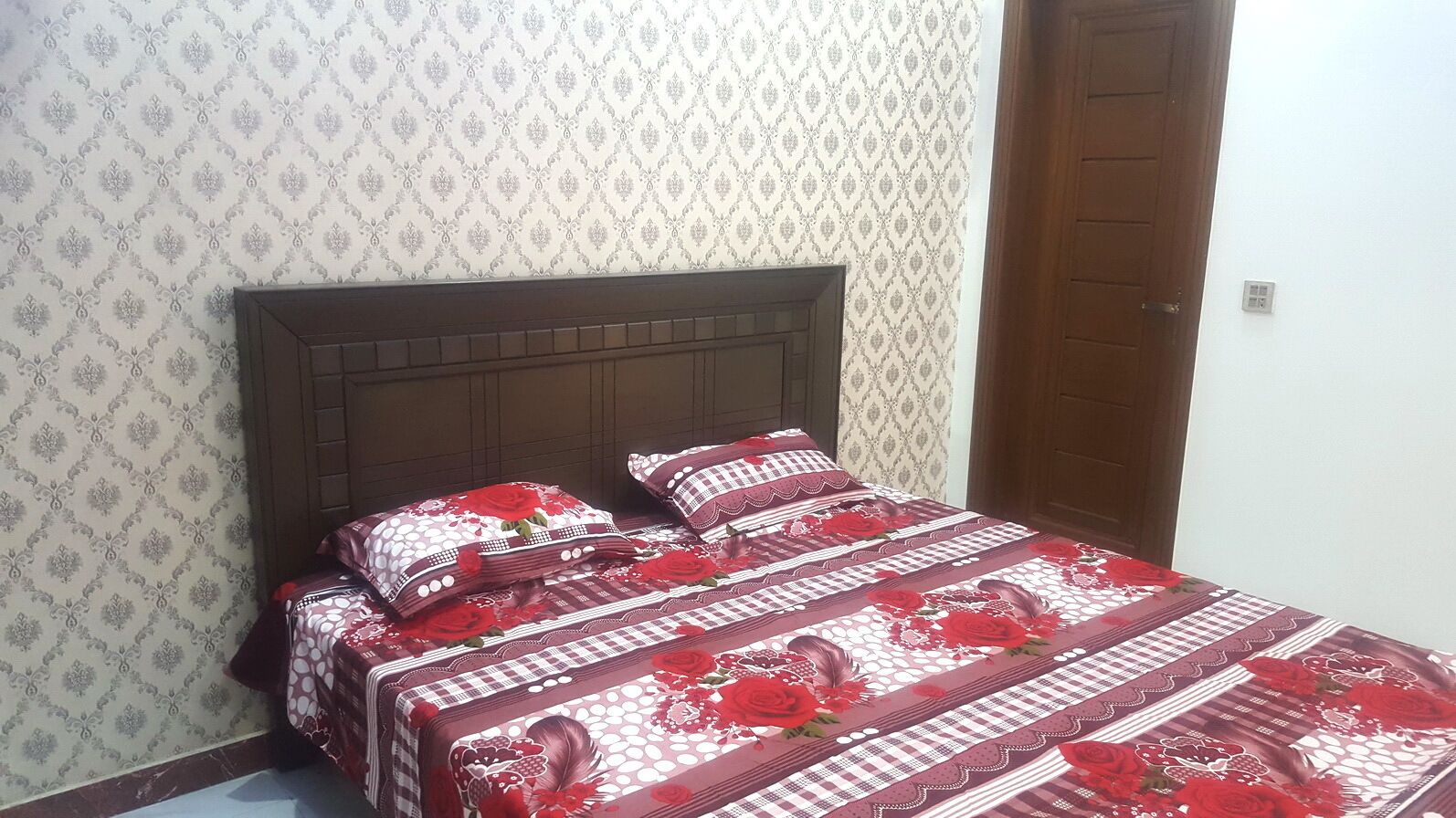 A bed with a floral-patterned bedspread and matching pillows, set against a patterned wallpaper and a wooden headboard.