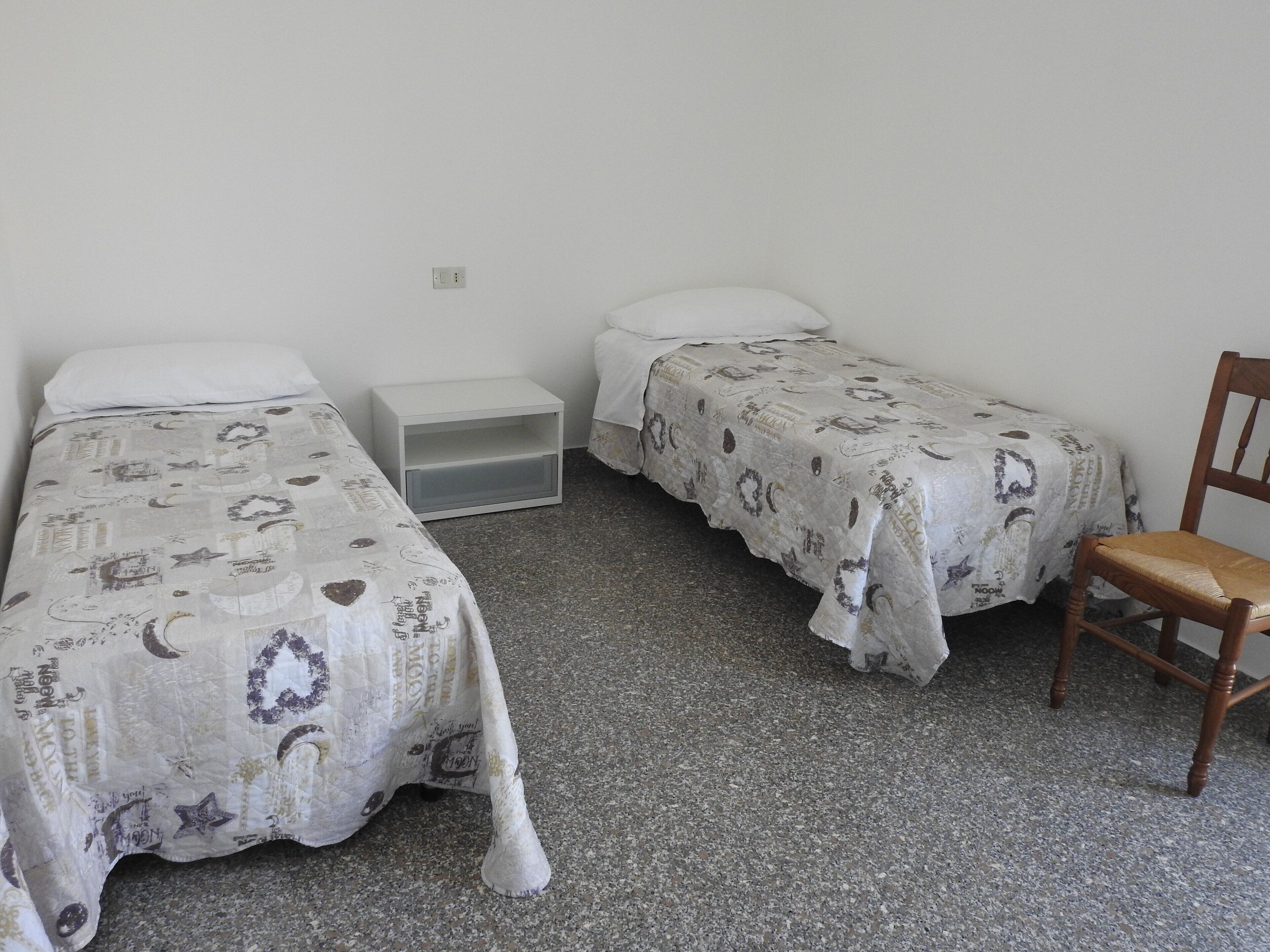 Ein kleines, einfach eingerichtetes Zimmer mit zwei Einzelbetten, einem Nachttisch und einem Stuhl.