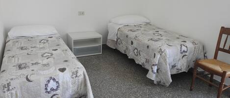 Ein kleines, einfach eingerichtetes Zimmer mit zwei Einzelbetten, einem Nachttisch und einem Stuhl.