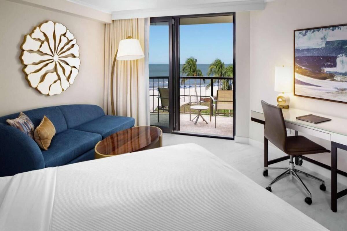 Een hotelkamer met een bed, een blauwe bank, een bureau met een stoel, een groot raam met uitzicht op het strand, en een decoratief wandobject.