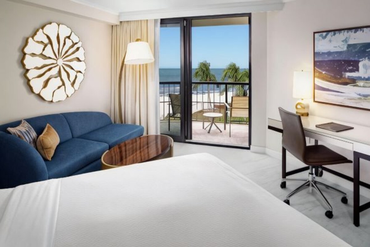 Una camera d'albergo con un letto, un divano blu, una scrivania, una sedia, un tavolino rotondo, un'ampia finestra vista mare e un elemento decorativo a parete.