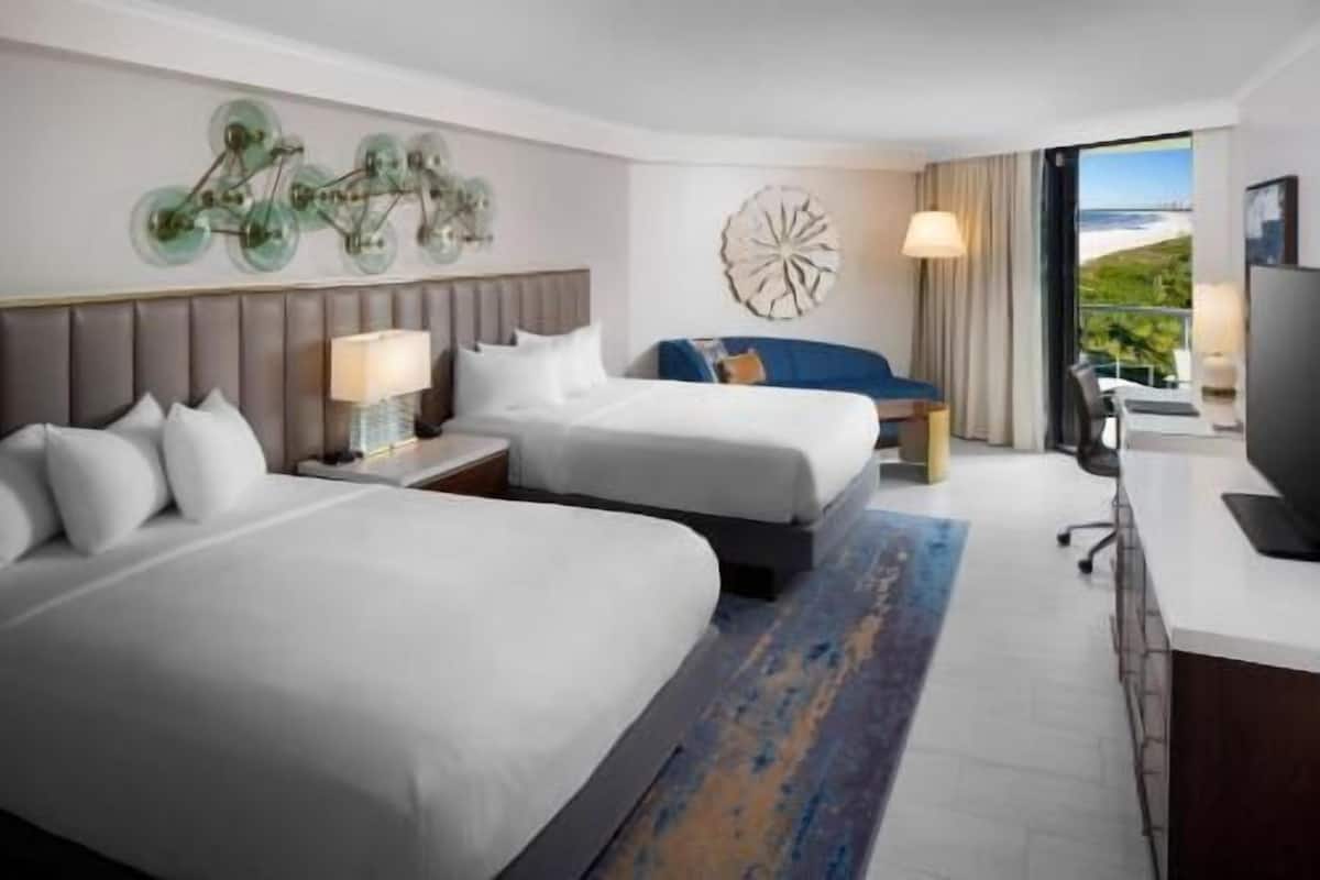 Hotelkamer met twee bedden, een bureau, een stoel, een lamp, een decoratief wandobject en een groot raam met uitzicht.