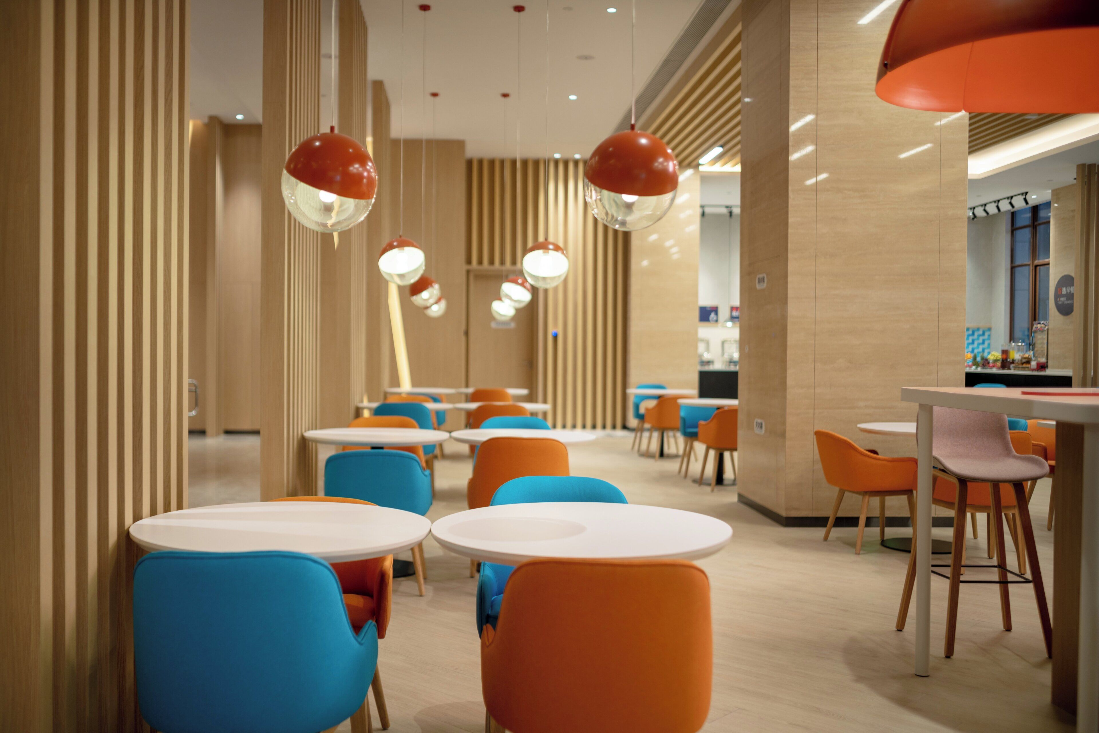 A modern interior with round tables and colorful chairs, a wooden floor, and pendant lighting.