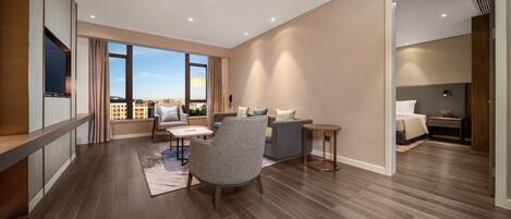 A modern living room with a sofa, armchair, and coffee table, large windows with a view, and a flat-screen TV mounted on the wall.