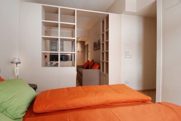 A bedroom with a bed, a nightstand, a lamp, and a wardrobe with open doors revealing a kitchen area.