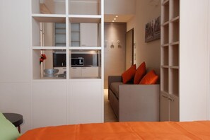 A modern bedroom with a bed, a grey sofa with orange cushions, a white wardrobe, and a kitchenette in the background. - UR-NEST Rizzoli 2 piano (Bologna)