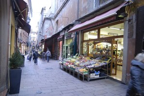 A narrow street with shops on either side, including a fruit and vegetable stall and a pharmacy. - UR-NEST Rizzoli 2 piano (Bologna)