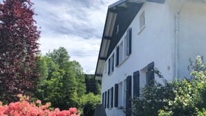 Un bâtiment blanc aux volets noirs, entouré d’une végétation luxuriante et de plantes en fleurs.