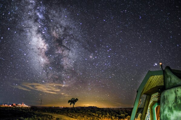 A starry night sky with the Milky Way visible, a lone tree, and a green structure with a dome-like roof.