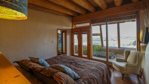 A bedroom with a large bed, a chair, and a view of the outdoors through large windows. - Spectacular Earthship (El Prado)