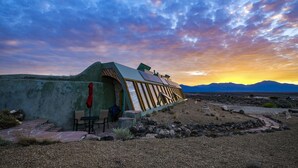 A modern building with a sloped roof, a small patio area with a table and chairs, and a pathway leading to the entrance. - Spectacular Earthship (El Prado)