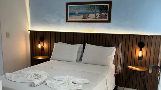 Un lit bien fait, avec du linge de lit et des serviettes blanches, une tête de lit en bois et deux tables de chevet fixées au mur, chacune avec une lampe.
