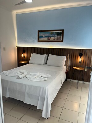 A neatly made bed with white linens and towels, a wooden headboard, and two wall-mounted nightstands with lamps. - Nascimento Suites (Búzios)
