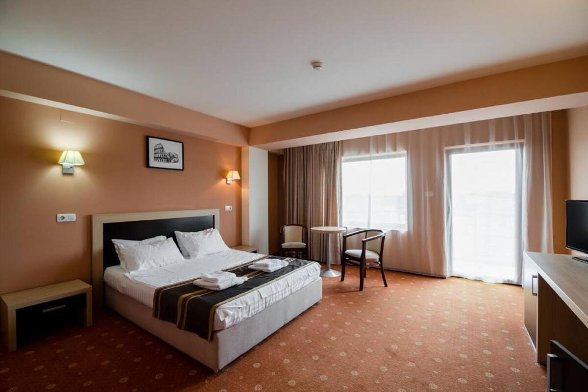 A hotel room with a bed, bedside tables, a desk, a chair, a television, and a window with curtains.