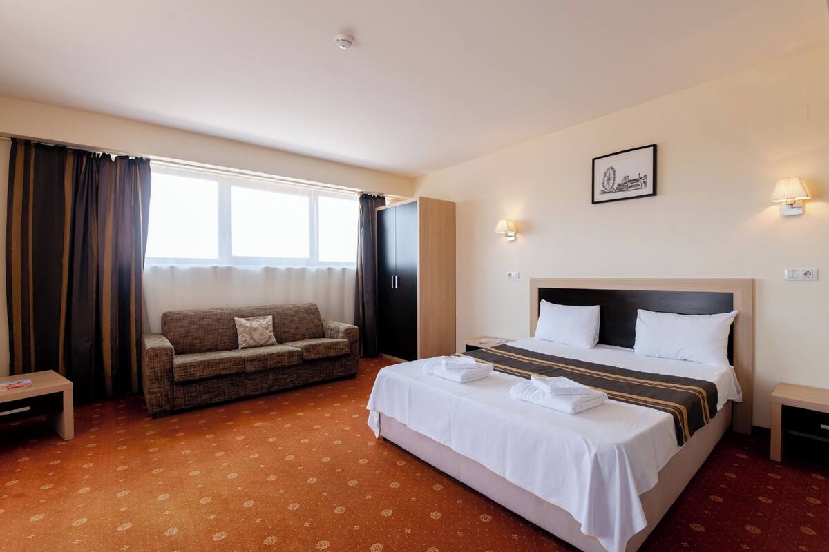 A hotel room with a large bed, a sofa, a small table, and a window with curtains.