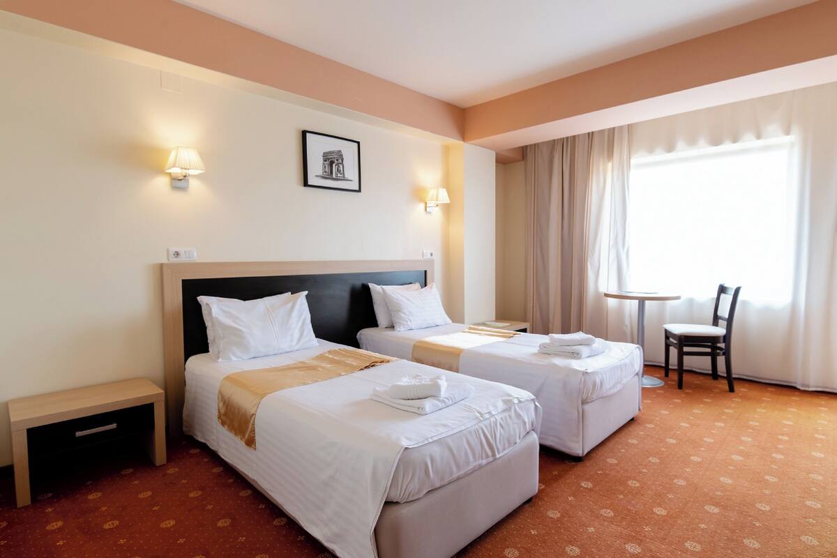 A hotel room with two beds, a small table, a chair, and a window with curtains.