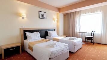 A hotel room with two beds, a small table, a chair, and a window with curtains.