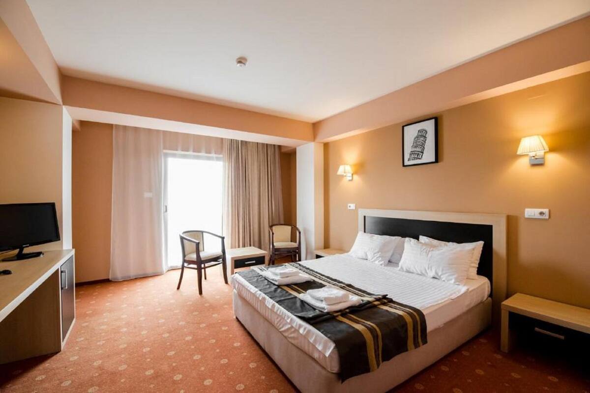 A hotel room with a bed, a desk with a computer, a chair, a lamp, and a window with curtains.