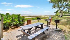 An outdoor seating area with picnic tables and barbecue grills, overlooking a beach. - Oceanfront Sunrises on Mitchelville Beach (Hilton Head Island)