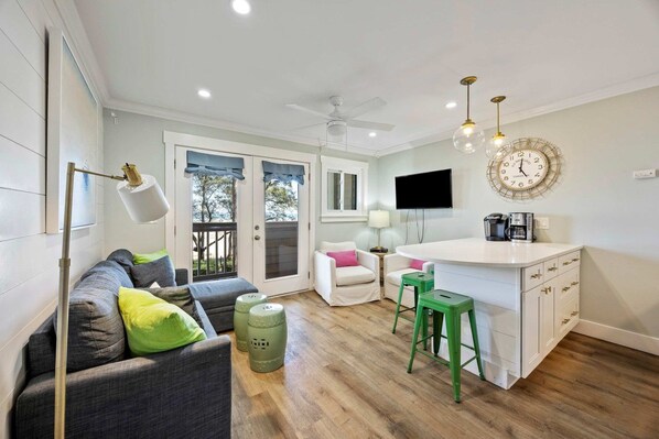 A modern living room with a sofa, green stools, a TV, and a kitchen area with a coffee machine.