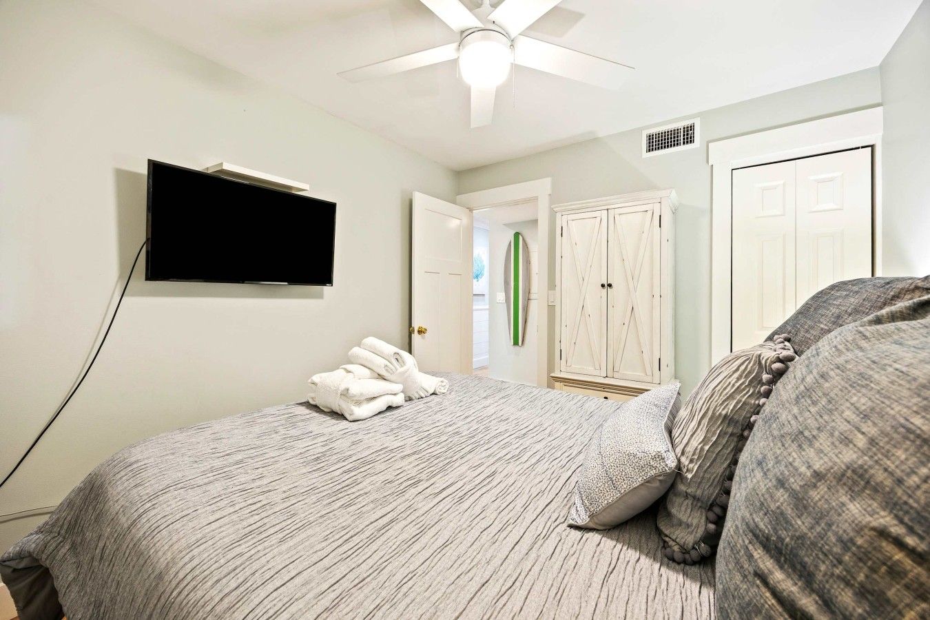 Un dormitorio con cama, televisor montado en la pared, ventilador de techo y puerta que da a otra habitación.
