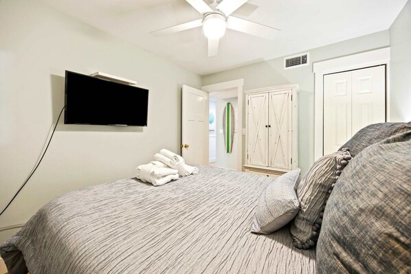 A bedroom with a bed, a television mounted on the wall, a ceiling fan, and a door leading to another room.