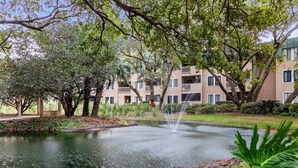 A tranquil courtyard with a pond, lush greenery, and a multi-story building with balconies. - Oceanfront Sunrises on Mitchelville Beach (Hilton Head Island)